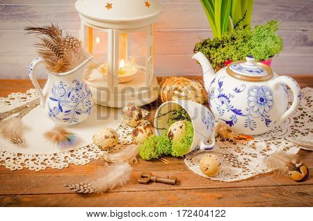 Spring floral still life with blue teapot, cup, moss. Lantern, feathers and quail eggs on wooden background