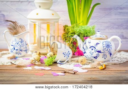 Spring floral still life with hyacinth, hearts. Blue teapot, cup, moss. Lantern, feathers and quail eggs on wooden background