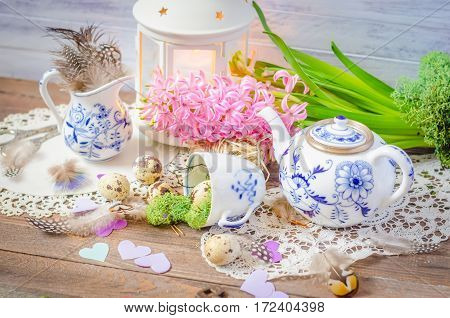 Spring floral still life with hyacinth, hearts. Blue teapot, cup, moss. Lantern, feathers and quail eggs on wooden background