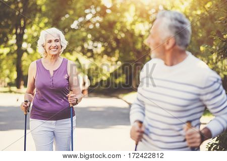 Sports activities. Joyful delighted aged woman holding the walking poles and looking at her husband while going after him