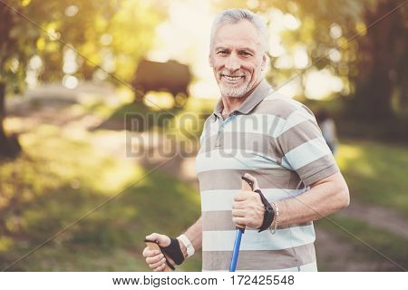 Sport walking. Delighted well built aged man smiling and enjoying his walk while being in the park