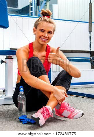 Woman in gym workout with fitness equipment. Girl drink from bottle water relaxing after workout sitting on floor at gym. Female tired after training and show thumb up.