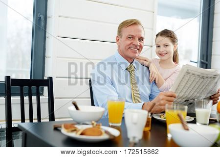 Businessman with newspaper and his daughter sitting by table in the kitchen