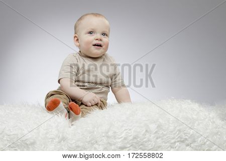 Adorable little baby sitting on the white blanket and looking up, studio shot, isolated on grey background.