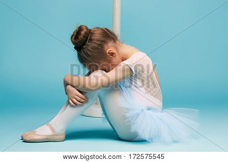 The little girl as balerina dancer sitting and posing near ballet rack on blue studio background