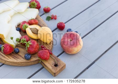 Wooden board with fresh organic fruit and berries: melon strawberries blueberries apricot peach and red current on grey background. Healthy snack