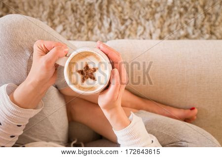 Girl with coffee mug sitting on the sofa indoors. Woman drinking a cup of coffee or tea sitting cozy at home. Relax and rest. Cappuccino with cinnamon star.