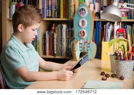 School kid boy sitting at his table and watching tablet pc at home indoors. Child with tablet computer in his room. Studying with tablet computer.