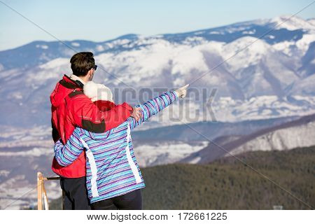 Rear view of a loving couple in fur hood jackets looking at snowed mountain range.
