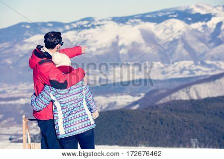 Rear view of a loving couple in fur hood jackets looking at snowed mountain range.