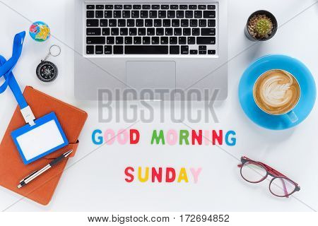 Caption word Good morning sunday. White office desk with laptop diary eyeglasses compass pen blank identification card and cup of coffee on white background.