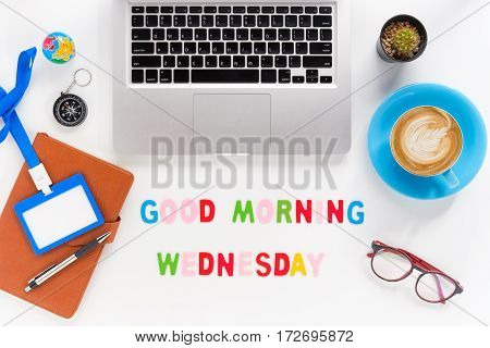 Caption word Good morning wednesday. White office desk with laptop diary eyeglasses compass pen blank identification card and cup of coffee on white background.