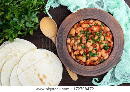White large beans in sweet and sour tomato sauce in a clay bowl on a dark background. The top view. Vegetarian cuisine. Lenten meal.