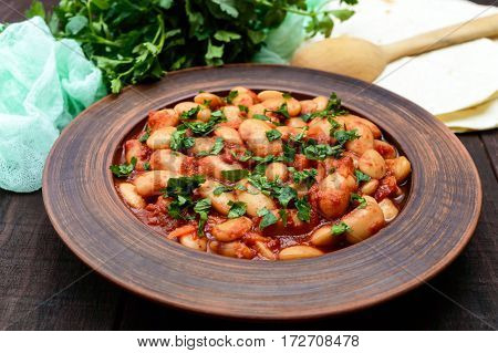 White large beans in sweet and sour tomato sauce in a clay bowl on a dark background. Vegetarian cuisine. Lenten meal.
