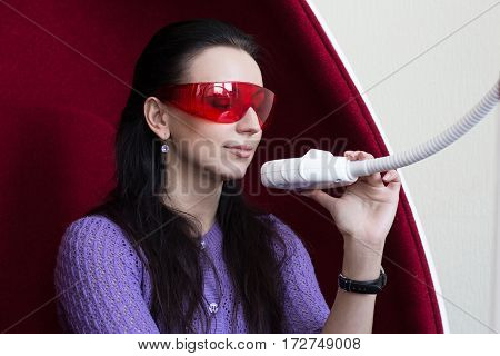 Young woman on a red high chair with red glasses holds dental aquipment of light in office