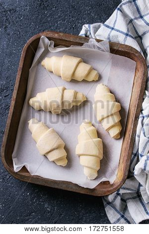 Raw unbaked croissant on baking paper in old oven tray with kitchen towel over black texture concrete background. Top view