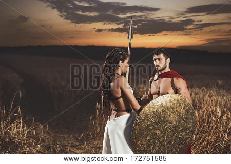 Portrait of couple meeting after war in field. Beautiful woman with long hair wearing like greece in white dress looking face to face with man warrior wearing like spartan.