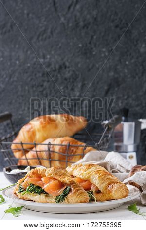 Croissant with smoked salted salmon, spinach and arugula served on white plate with bowl of cream cheese and coffee maker over white and gray concrete background. Copy space