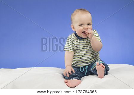 Laughing 8-month-old baby boy, sitting on the white blanket , studio shot, isolated on blue background.