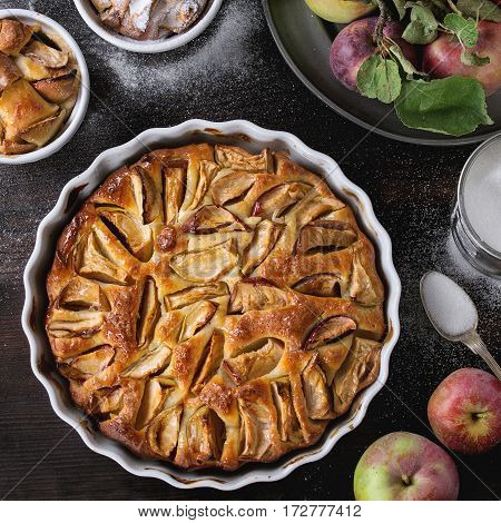 Homemade big and small apple cake pie in white ceramic forms with fresh apples with leaves, sugar powder, and tin can of sugar over dark wooden background. Top view