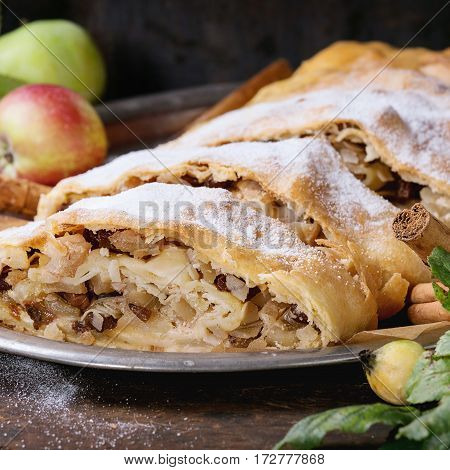 Sliced homemade apple strudel served with fresh apples with leaves, cinnamon sticks and sugar powder on vintage metal tray over old wooden background. Close up, dark rustic style