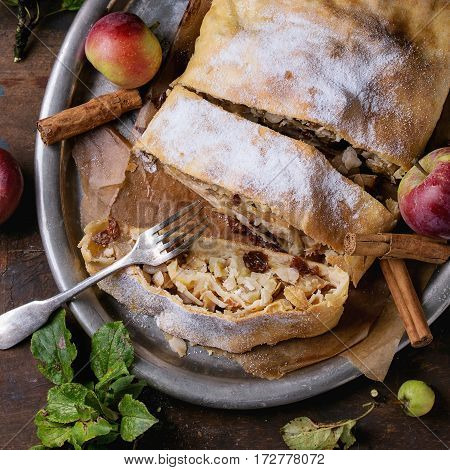 Sliced homemade apple strudel served with fresh apples with leaves, cinnamon sticks and sugar powder on vintage metal tray with fork over old wooden background. Overhead view, rustic style