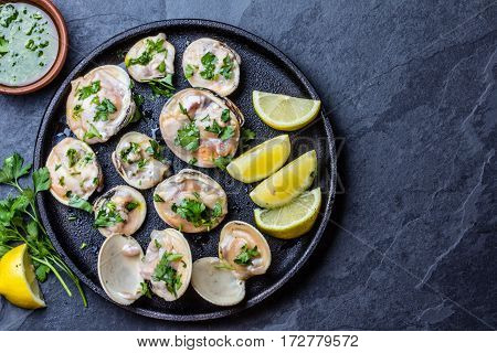 Fresh raw clams with lemon, herbs and white wine on blackiron plate on stone slate background. Top view. Free space for text