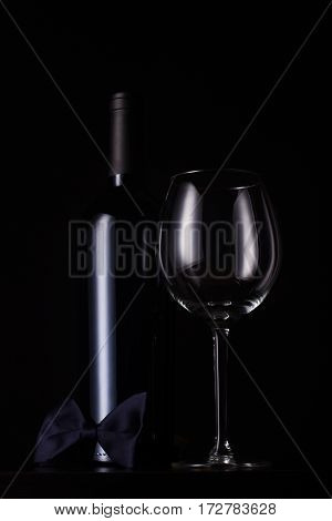 vertical front view of black red wine bottle with cap and no label tall empty glass and a dark bow tie at the base