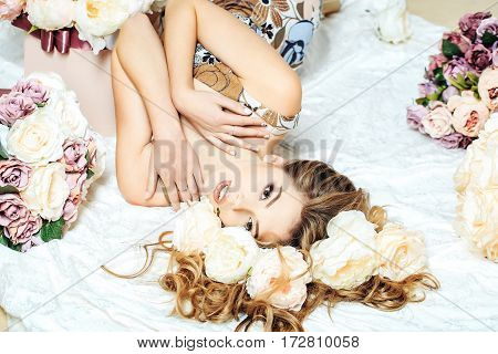 Sexy young pretty woman or cute girl with long hair in floral dress laying among lilac rose and peony flowers on white bedsheet