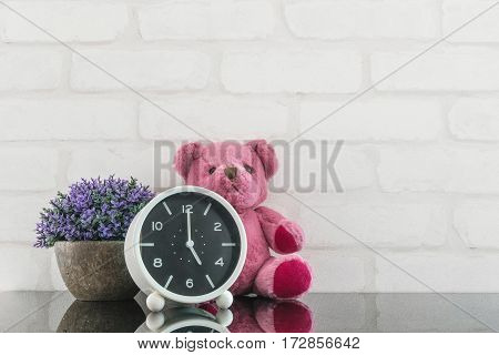 Closeup black and white alarm clock for decorate in 5 o'clock with bear doll and plant on black glass table and white brick wall textured background with copy space