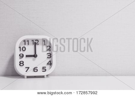 Closeup white clock for decorate in 9 o'clock on white wood desk and cream wallpaper textured background in black and white tone with copy space