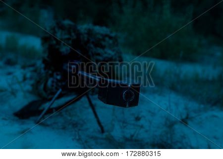 Army sniper with big rifle lying in wait in the forest at nighttime