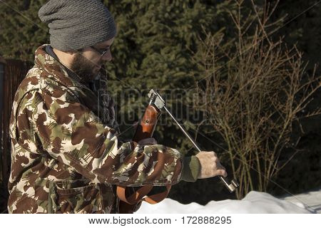 Man in winter forest reloads pneumatic weapons. 
Hunter dressed in camouflage with pneumatic gun, rifle 
The man in the white winter forest loads long arms.