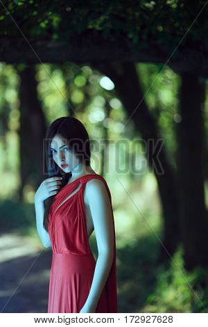 Wonderful sexy fashion model walking in a fantastical forest by footpath.Mystical portrait of sensual seductive woman in luxury long red dress posing against bokeh trees background. Multi-racial Asian Caucasian girl. Fashionable toning. Creative computer 