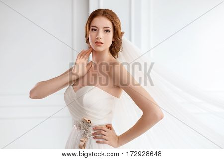 Bride in white dress and long veil indoors. Young gorgeous fashion model posing in white interior studio's room near big windon. Portrait shot