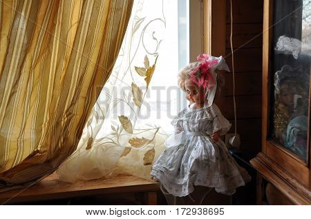 Big doll in a smart white dress and with a bow in her hair on a wooden window sill. Russia
