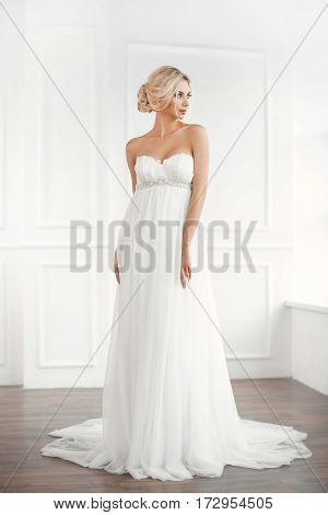 Classical young gourgeous bride. Studio interior fashion shot of fashion model in wedding dress in white room. Blonde woman in full lenght looking away.