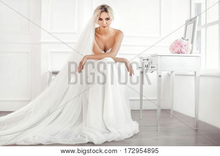 Classical young gourgeous bride. Studio interior fashion shot of fashion model in wedding dress with bouquet of flowers sitting in white room with chair bedside table and mirror. Blonde woman portait with profeshional make-up and hairstyle.