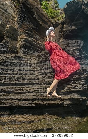 A Young Woman In A Red Dress And White Hat Stands On A Rock By The Sea And Holds A Hat With Her Hand
