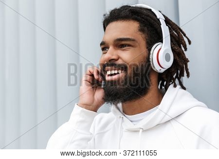 Photo of a handsome optimistic cheery young man walking outdoors while listening music by headphones