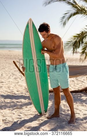 Caucasian man enjoying his time at the beach with his friends on a sunny day, standing shirtless, holding a surfboard and looking at it