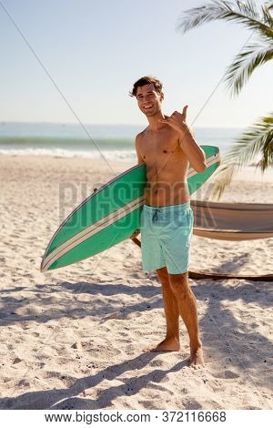 Caucasian man enjoying his time at the beach with his friends on a sunny day, standing shirtless, holding a surfboard and looking at the camera