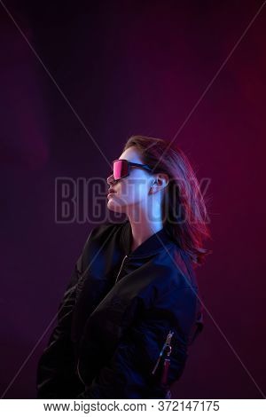 Portrait Of A Cool Girl In Black Bomber Jacket With Sunglasses On A Dark Background In Neon Light.