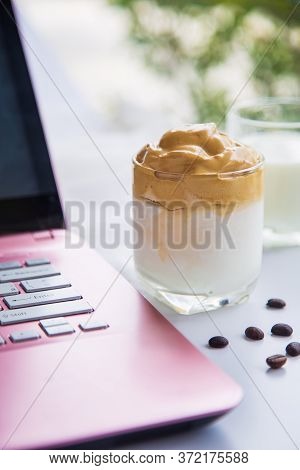 Close Up A Cup Of Dalgona Coffee With Coffee Beans Next To A Pink Laptop On A White Table With A Cup