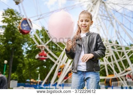 Portrait Of A Child With Sweet Cotton Candy. A Little Girl On The Background Of Ferris Wheel Is Eati