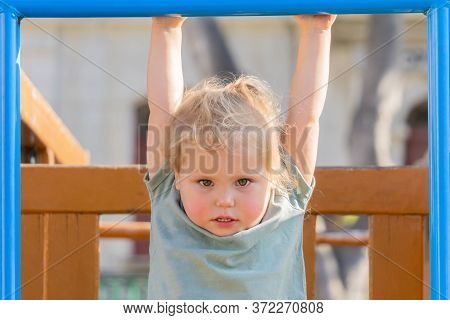 Little Girl Playing At Playground And Hanging On Crossbar. Todler Hanging On Horizontal Bar In Playg