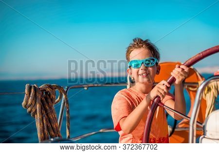 A Handsome Boy Of 9-10 Years Old At The Helm Of A Yacht In Bright Orange Clothes, Blue Sunglasses Ag