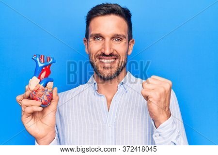 Young handsome man holding heart organ with veins and arteries over blule background screaming proud, celebrating victory and success very excited with raised arm