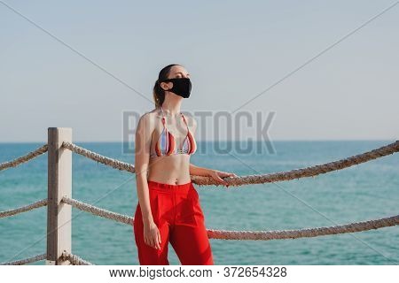 European Woman In A Bikini Top And Red Pants Wearing Protective Face Mask At The Public Beach. New N