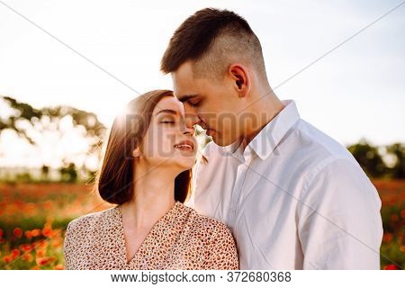 Young Romantic Couple Standing On The Red Poppy Field On A Sunny Day Showing Love To Each Other. Hap